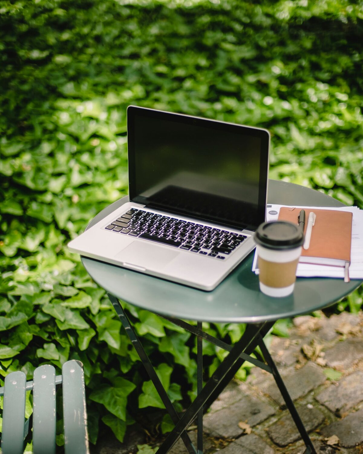Laptop, to-go coffee and a notebook on a small round table outdoors, in front of green ivy